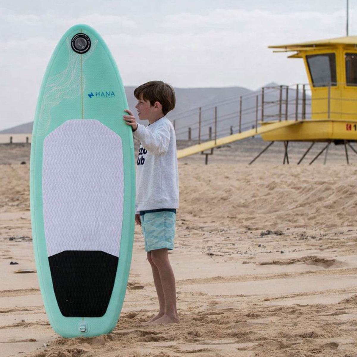 Enfant à côté de la planche de surf gonflable HANA Premium 5'4, sur la plage