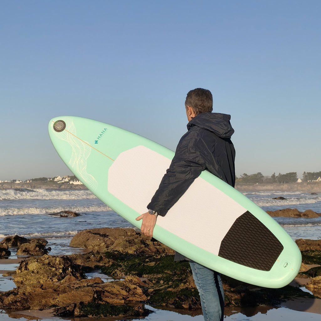 Surfeur portant la planche de surf gonflable HANA Premium 6'6 au bord de l’eau