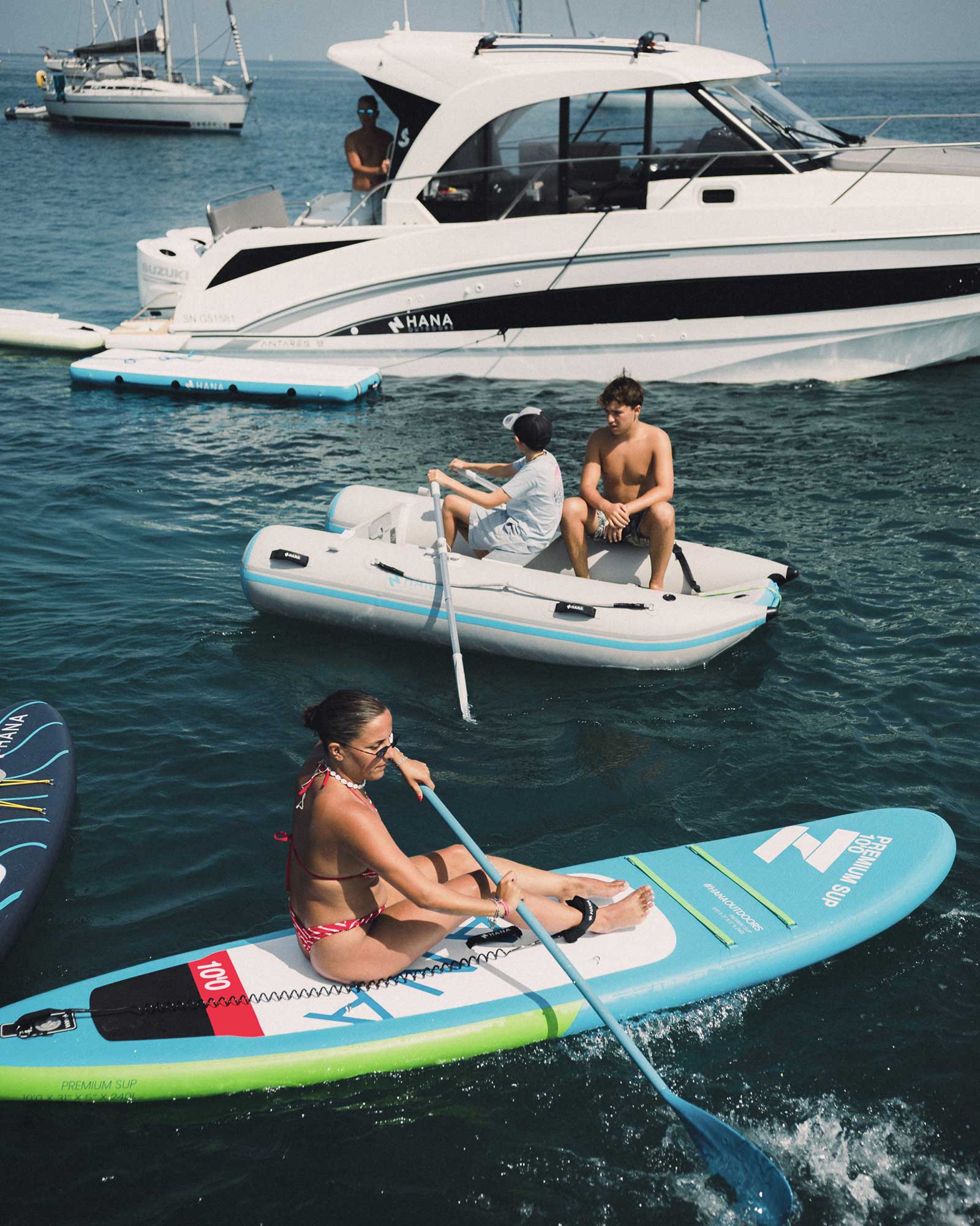 At sea around a HANA boat: a woman on a paddle board and two youngsters in an inflatable tender